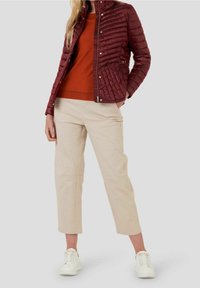 Veste matelassée bordeaux avec boutons-pression, associée à un pull orange et un pantalon beige. Des baskets blanches complètent la tenue.