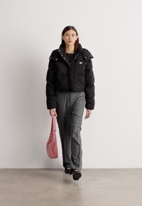 Casaco de puffer preto curto, com costura horizontal e botões de pressão. Calças cinza risca de giz e uma bolsa rosa completam o conjunto.