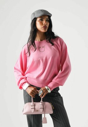 Donna con berretto grigio e maglione rosa che tiene una piccola borsa rosa, in piedi davanti a uno sfondo chiaro e uniforme.