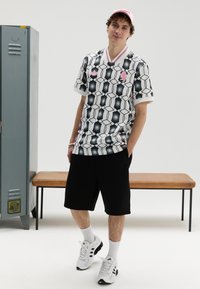 Short-sleeve, white shirt with a geometric black pattern, pink accents, and a collar. Worn with black shorts and white sneakers.