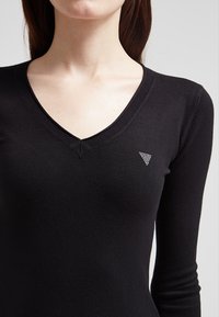 Černý svetr s dlouhým rukávem a výstřihem do V z měkkého pleteného materiálu, se malým trojúhelníkovým akcentem s diamantovým vzorem na levé hrudi.