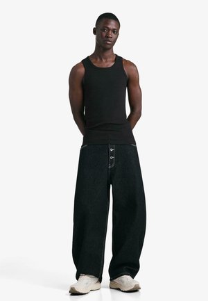 Crna pamučna majica bez rukava s hlačama širokih nogavica od tamnog trapera s tri gumba i kontrastnim prošivima. Obuća su bež tenisice.