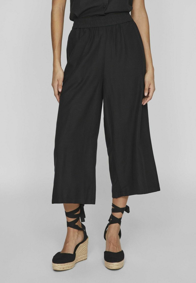 Pantalones anchos negros con cintura elástica, hechos de una tela ligera, combinados con zapatos de esparto negros con tiras entrecruzadas.