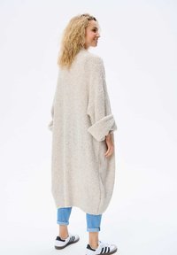 Overmaat romige, gestructureerde gebreide cardigan met opgerolde mouwen, gecombineerd met lichtblauwe jeans en witte sneakers met zwarte accenten.