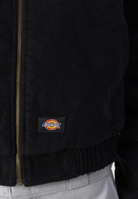 Giacca in corduroy nera con zip frontale, polsini a coste e orlo. Presenta una patch con il logo Dickies sul lato sinistro del petto.