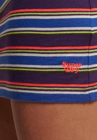 Streifenstoff in Blau, Lila, Grün, Weiß und Orange auf Kleidung mit gesticktem roten "Super Dry"-Text nahe dem Saum über dunkler Haut.