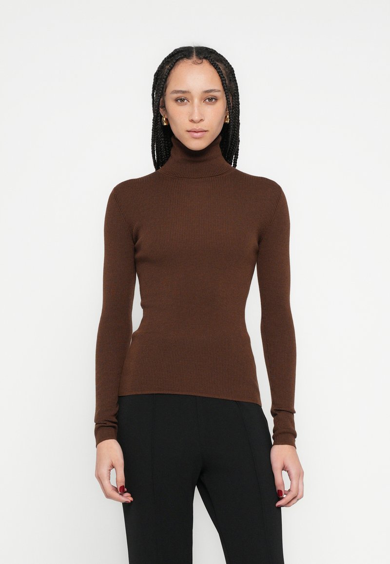 Pull à col roulé marron côtelé à manches longues, coupe ajustée, texture douce, associé à un pantalon noir, fond minimaliste.