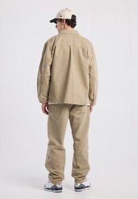 Giacca di jeans beige con vestibilità rilassata, abbinata a pantaloni beige larghi e sneakers bianche con accenti grigi. Cappellino a righe bianche e nere.