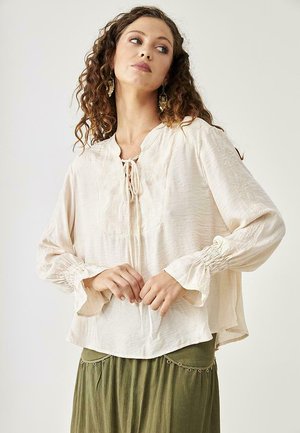Blusa ligera color beige claro, fluida, con escote redondo, detalle de lazo y acentos bordados. Mangas largas con puños fruncidos. Tejido texturizado.