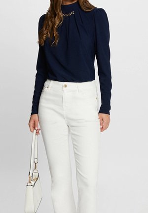 Frau trägt ein marineblaues Langarmoberteil mit Kettendetail, weiße High-Waist-Jeans und hält eine kleine weiße Handtasche an ihrer Seite.