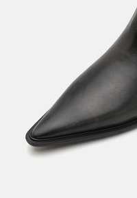 Crne kožne čizme s visokom petom i šiljastim prstom, glatke teksture i vitke siluete. Ima minimalistički dizajn i elegantan profil.
