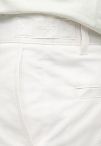 Gros plan d'un pantalon blanc avec une étiquette de ceinture cousue embossée "HUGO" et une poche arrière fendue.