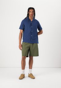 Blue short-sleeve shirt with white buttons, paired with olive green shorts. The model wears light brown sneakers and white socks.