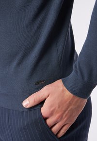 Marineblauer Strickpullover mit gerippten Bündchen und Saum, versehen mit einem kleinen schwarzen Logo-Tag. Glatte Textur, figurschmeichelndes Design, kombiniert mit gestreiften Hosen.