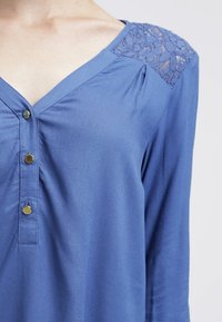 Blue blouse with a V-neck, three gold buttons, pleats at the neckline, and lace accents on the shoulders. Soft fabric with a smooth texture.