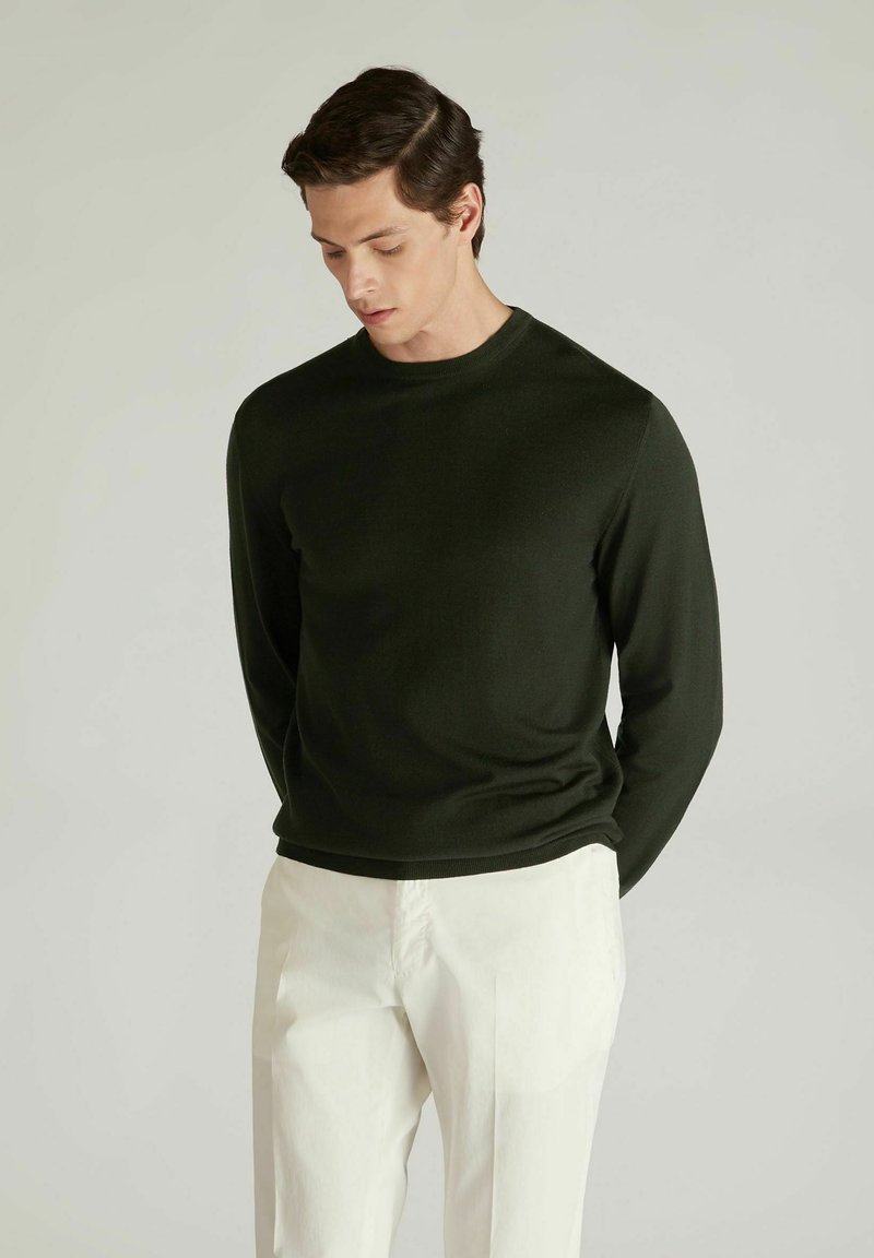 Dunkelgrüner Pullover mit langen Ärmeln und rundem Ausschnitt, aus glattem Strickstoff gefertigt. Kombiniert mit weißen Hosen, die eine schlanke Passform zeigen.