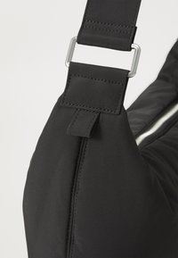 Esquina de una bolsa de nylon negra con herrajes plateados, que muestra una textura acolchada, correa ajustable y una apertura con cremallera a lo largo de un lado.