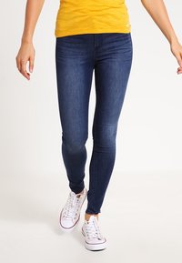 Úzké džíny z elastického denimu, tmavě modré barvy, přiléhavý střih, v kombinaci s bílými teniskami, nošeny s žlutým topem.