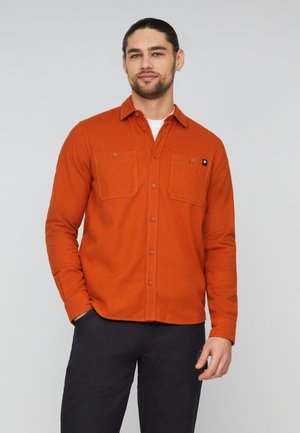 Mand med mørkt hår og skæg iført en orange skjorte med knapper over en hvid t-shirt og mørke bukser, stående mod en ensfarvet lys baggrund.