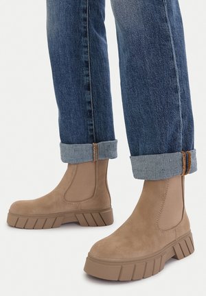 Botines beige de ante con suela de goma gruesa, con paneles elásticos en los lados, combinados con jeans azul oscuro remangados.