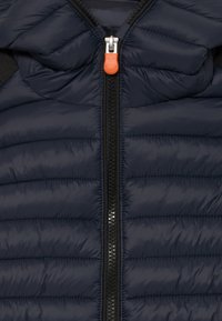 Vesta puffer de culoare bleumarin, cu fermoar complet și o curea portocalie și accente negre. Textură căptușită cu model matlasat și glugă.