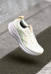 White athletic shoe with green accents, a textured upper, and a sleek curved sole. Features laces and a light tan heel tab.