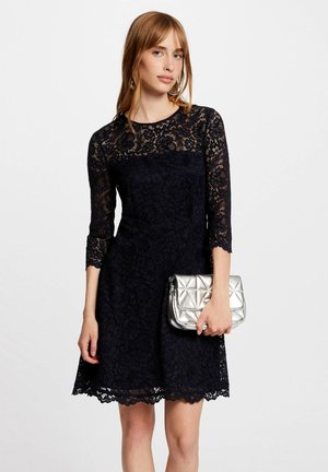 Navy lace dress with three-quarter sleeves, scalloped hem, and flower pattern; paired with a metallic silver quilted handbag.