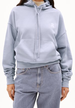 Persoon draagt een lichtblauwe cropped hoodie met voorzak en trekkoorden, gecombineerd met lichtblauwe jeans met hoge taille, staand met ontspannen handen.