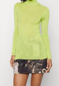 Langärmliges Turtleneck-Kleid in leuchtendem Limettengrün mit sheer Textur, kombiniert mit einem gemusterten schwarzen Minirock mit abstrakten Designs.