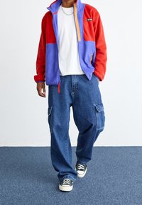 Chaqueta de forro polar roja y púrpura con detalles en naranja, camiseta blanca debajo y pantalones cargo azul. Zapatillas negras y blancas completan el look.