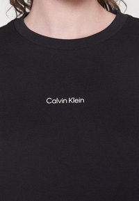 T-shirt nera in cotone, con design a girocollo. Un logo bianco "Calvin Klein" è posizionato al centro del davanti. Texture liscia.