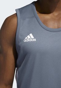 Šedý sportovní tílko vyrobené z prodyšné tkaniny. Má kulatý výstřih a bílé logo Adidas na hrudi. Vertikální švy.