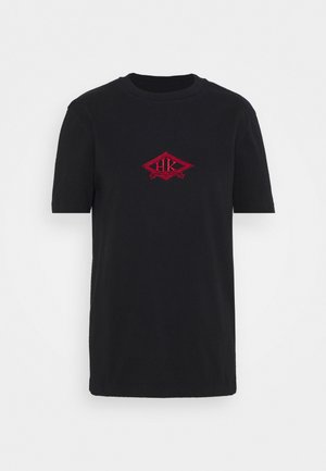 Musta puuvillainen t-paita, jossa on lyhyet hihat ja pyöreä pääntie. Rinnan kohdalla on punainen kirjailtu timantin muotoinen logo.