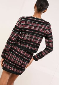 Černý, červený a bílý kostkovaný krátký svetr s texturovanou přízí, s dlouhými rukávy a přiléhavým střihem, nošený s odpovídající sukní.