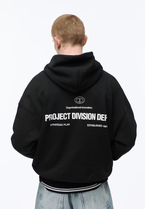 Oseba kratkih las, videna od zadaj, oblečena v črno kapuco z belim napisom "PROJECT DIVISION DEP" in svetlo modre kavbojke.