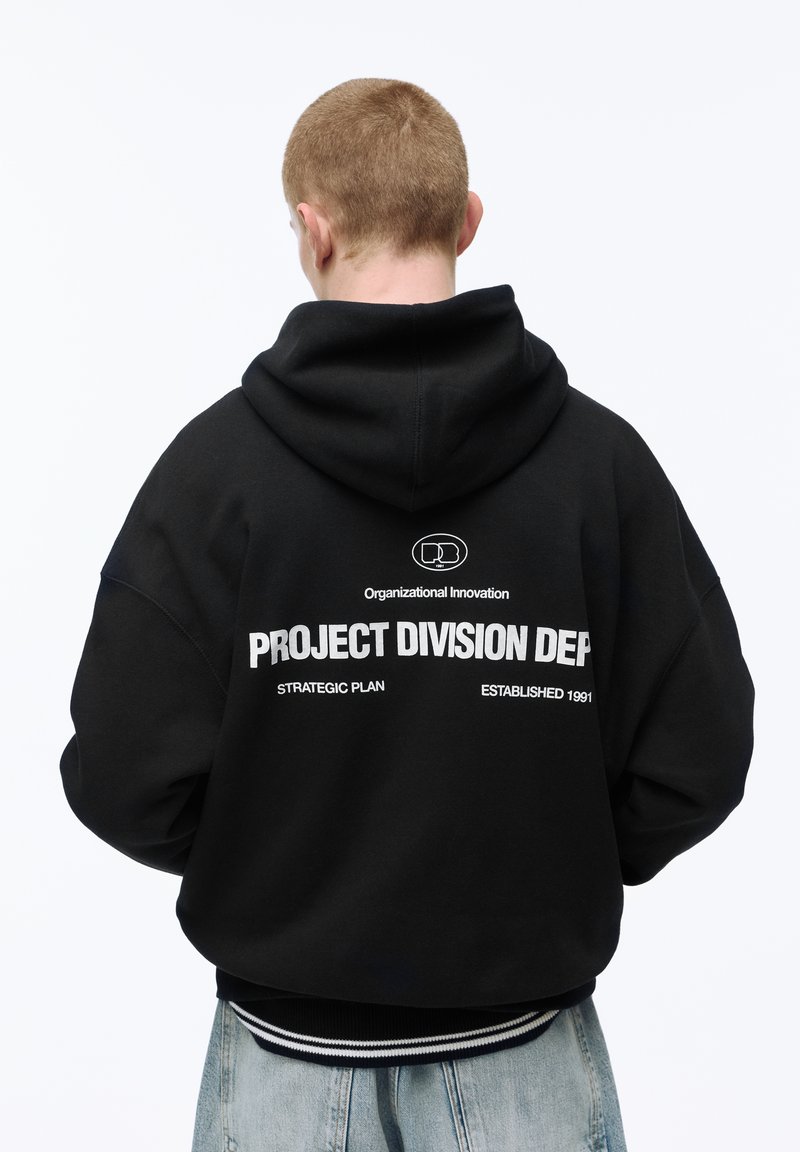 Person mit kurzen Haaren, von hinten gesehen, trägt einen schwarzen Hoodie mit weißem Text "PROJECT DIVISION DEP" und hellblaue Jeans.