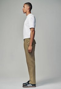 Maglietta bianca a maniche corte abbinata a pantaloni beige e scarpe da ginnastica nere con strisce bianche. Modello mostrato in vista di profilo.