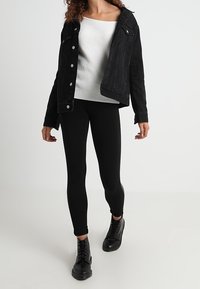 Giacca di jeans nera con bottoni, top bianco a costine con spalle scoperte, leggings neri e stivali neri con lacci. Silhouette semplice e aderente.