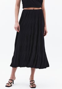Zwarte geplooide midi-jurk van textuurstof, met een hoge taille en een vloeiende zoom. Gepaard met zwarte sandalen met banden.