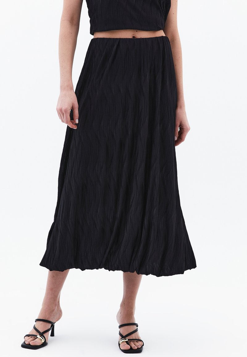 Zwarte geplooide midi-jurk van textuurstof, met een hoge taille en een vloeiende zoom. Gepaard met zwarte sandalen met banden.