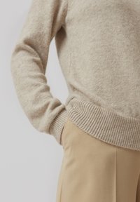 Beige strikket sweater med ribbet kant og manchetter, parret med lysebeige bukser, der viser en glat tekstur og en afslappet pasform.