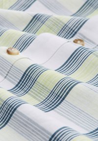 Katoenen stof met een golvend patroon in wit, lichtgroen en blauw. Bevat een houten knoopdetail bij de naad. Gestructureerde uitstraling.