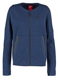 Marinblå zip-up-jacka gjord av mjukt, strukturerat tyg. Har två framfickor med grå accenter och rund halsringning. Nike-logotyp visas.