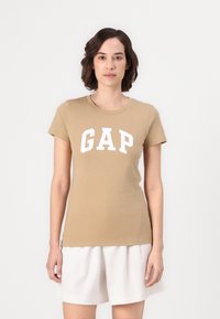 Beige cotton t-shirt with short sleeves and a crew neck, featuring bold white "GAP" lettering across the chest. Soft, smooth texture.
