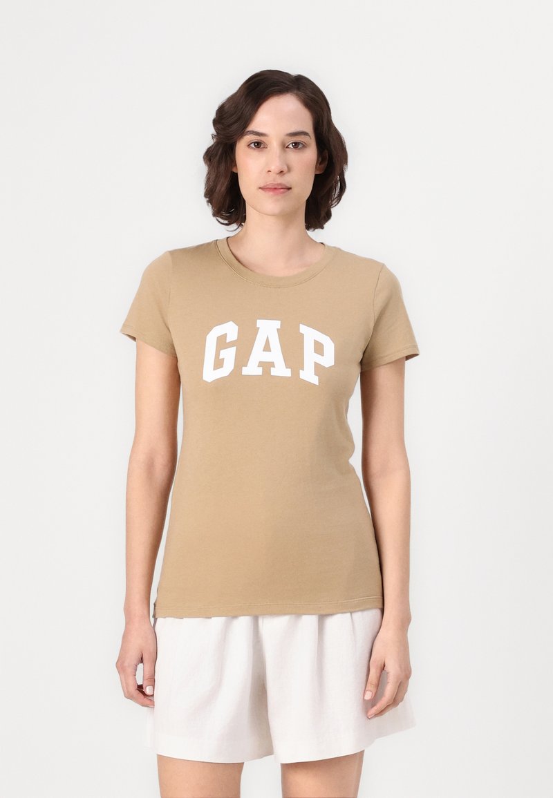 Beige cotton t-shirt with short sleeves and a crew neck, featuring bold white "GAP" lettering across the chest. Soft, smooth texture.