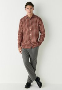 Jeune homme adossé à un mur, portant une chemise à carreaux rouge, un pantalon gris et des chaussures noires, avec une main dans la poche.