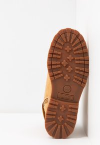 Hnědá gumová podrážka s výraznými vzory pro záběr, s logem Timberland. Horní část je vyrobena z béžové kůže s detaily prošití.