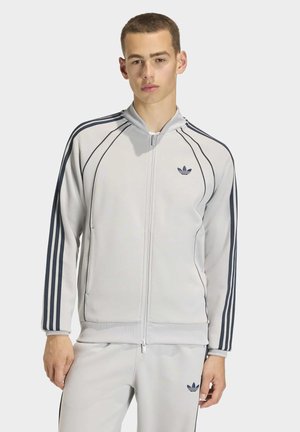 Young man wearing light gray Adidas tracksuit with black stripes on sleeves and pants, standing against plain background.