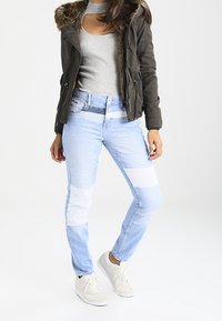 Jasnoniebieskie dżinsy o wąskim kroju w patchworkowym stylu z białymi łatami, w połączeniu z szarym, prążkowanym topem i ciemnozieloną kurtką z futrzanym wykończeniem. Białe sneakersy.