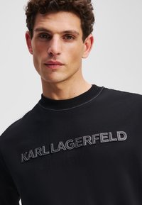 Černá mikina s výšivkou "KARL LAGERFELD" v bílé nitě. Kulatý výstřih s ribbovaným lemem a hladkou texturou.